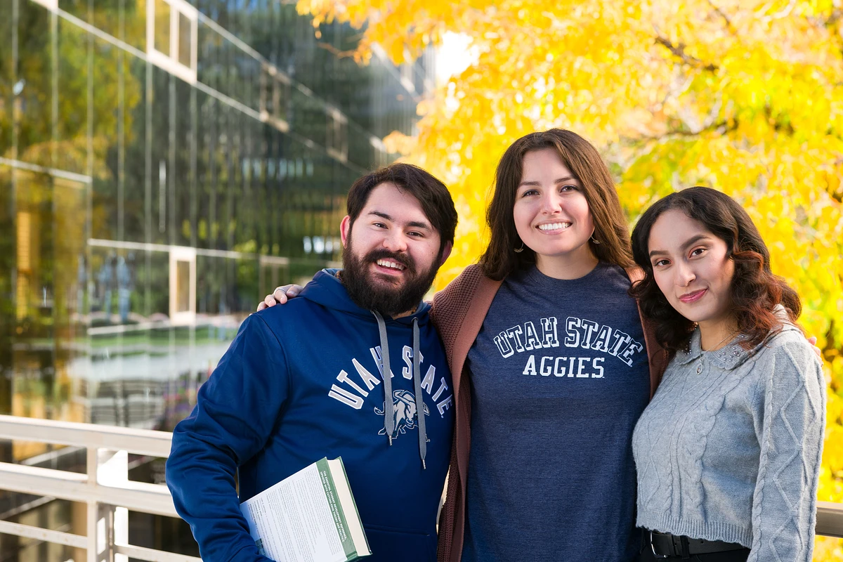 Three people are smiling and posing together outdoors, with one holding a book and wearing Utah State apparel.
